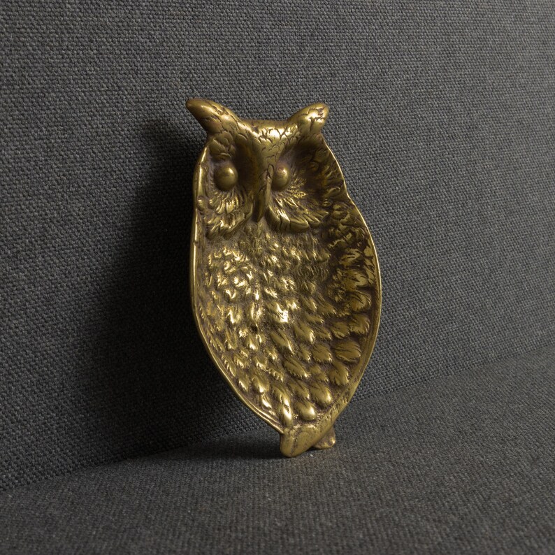 May include: A brass owl-shaped dish featuring a detailed, textured feather pattern across its surface. The metal tray has a distinct, rounded owl head at the top with two prominent circular eyes and a pointed beak, transitioning into a tapered body.