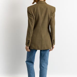 May include: A tan and black houndstooth blazer with three buttons on each cuff. The blazer is paired with blue jeans and brown sandals. The model is facing away from the camera.