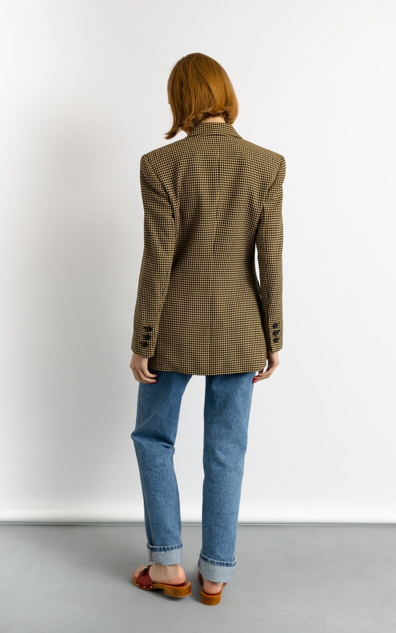 May include: A tan and black houndstooth blazer with three buttons on each cuff. The blazer is paired with blue jeans and brown sandals. The model is facing away from the camera.