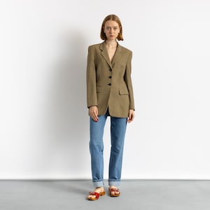 May include: A woman wearing a brown and black houndstooth blazer, blue jeans, and red sandals. The blazer has a classic tailored design with black buttons. The jeans are a light blue wash.