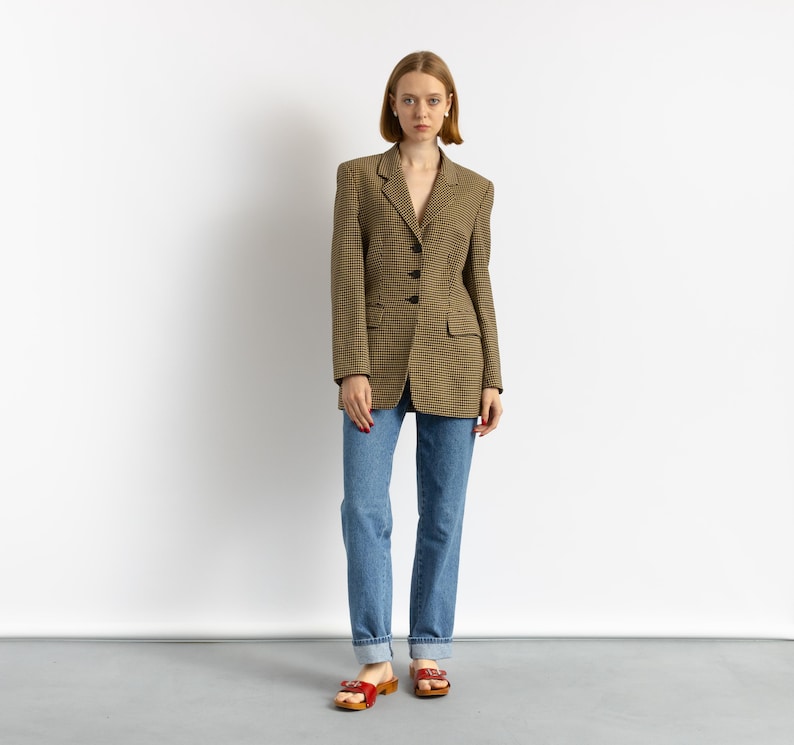 May include: A woman wearing a brown and black houndstooth blazer, blue jeans, and red sandals. The blazer has a classic tailored design with black buttons. The jeans are a light blue wash.