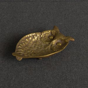 May include: A small, shallow brass dish shaped like an owl. The metal surface features embossed feather textures across the body and wings, with two smooth, raised circular eyes. The dish has a metallic gold finish and a curved, concave form.