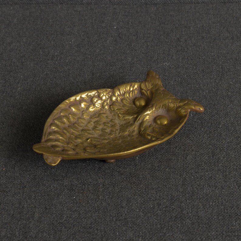 May include: A small, shallow brass dish shaped like an owl. The metal surface features embossed feather textures across the body and wings, with two smooth, raised circular eyes. The dish has a metallic gold finish and a curved, concave form.