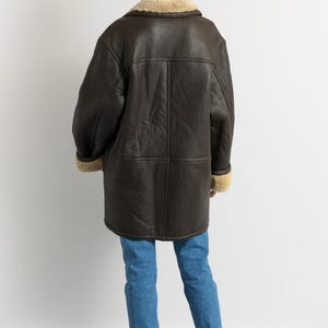 May include: A dark brown leather coat with a shearling collar and cuffs. The coat is worn over blue jeans and red boots. The coat has a classic, vintage style.