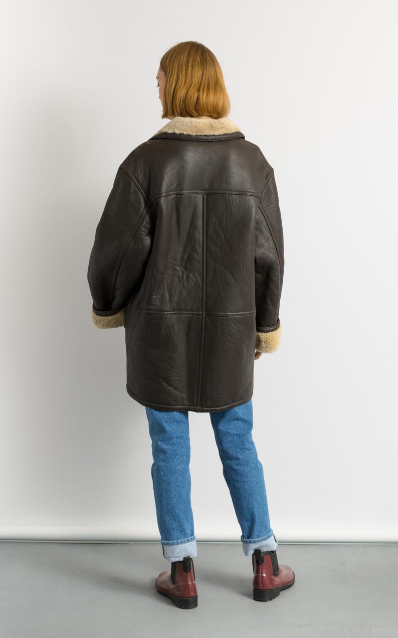 May include: A dark brown leather coat with a shearling collar and cuffs. The coat is worn over blue jeans and red boots. The coat has a classic, vintage style.