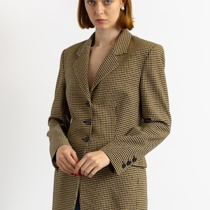 May include: A houndstooth blazer in black and tan. The blazer has a notched lapel, three-button closure, and three buttons on each cuff. The model is wearing blue jeans. The blazer is a classic style.
