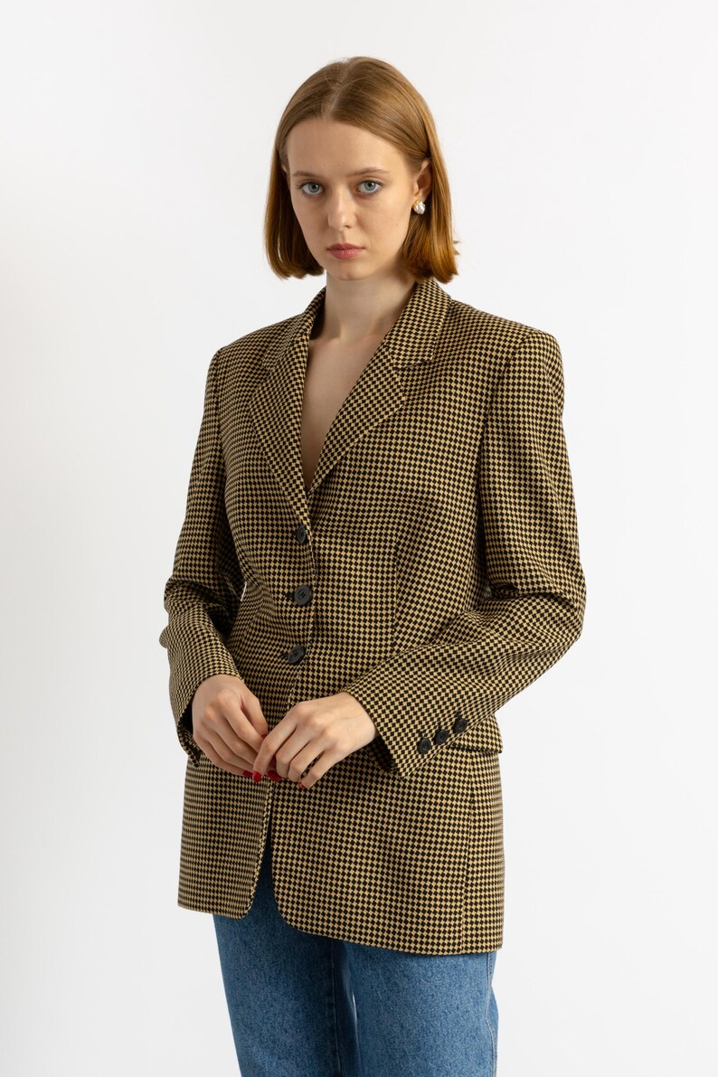 May include: A houndstooth blazer in black and tan. The blazer has a notched lapel, three-button closure, and three buttons on each cuff. The model is wearing blue jeans. The blazer is a classic style.