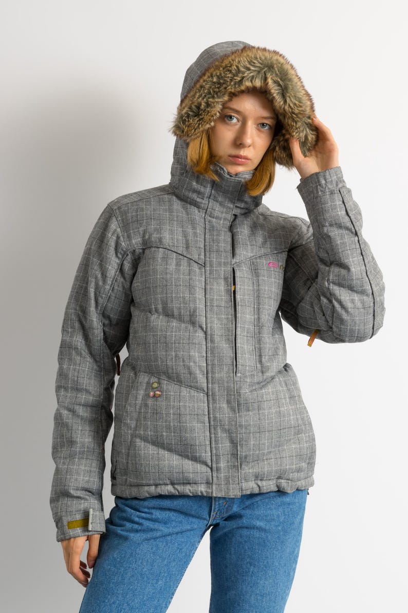 Puede incluir: Una chaqueta de invierno gris a cuadros con capucha forrada de piel sint&eacute;tica. La chaqueta tiene un dise&ntilde;o acolchado y un cuello alto. Cuenta con cierre de cremallera y bolsillos. El modelo lleva vaqueros azules.