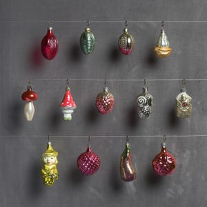 Puede incluir: Una colección de adornos navideños vintage en varias formas y colores, incluyendo rojo, verde, plateado y dorado. Los adornos están suspendidos de finos alambres contra un fondo gris. La colección incluye adornos con forma de hongo, fruta y personajes.