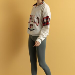 May include: A woman wearing a white sweatshirt with a red and black plaid sleeve and the text "SAN FRANCISCO" on the front. She is also wearing gray leggings and red sandals.