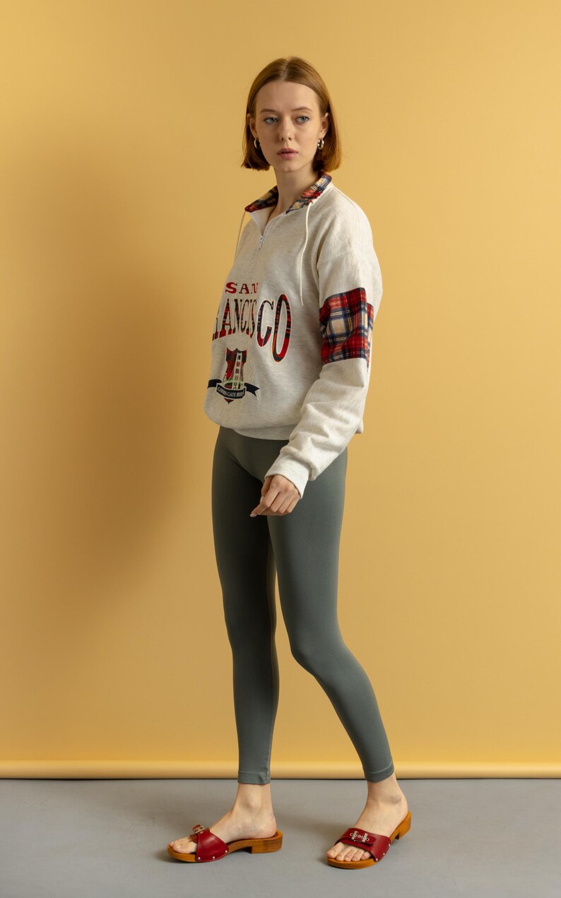 May include: A woman wearing a white sweatshirt with a red and black plaid sleeve and the text "SAN FRANCISCO" on the front. She is also wearing gray leggings and red sandals.