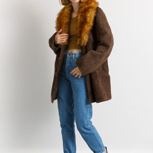 May include: A brown suede coat with a faux fur collar. The coat is worn with blue jeans and burgundy ankle boots. The model is standing in front of a white background.