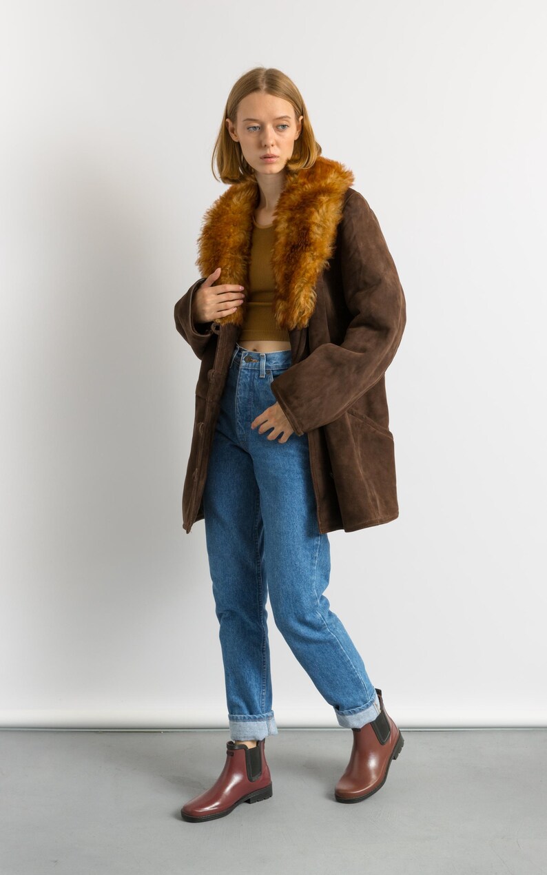 May include: A brown suede coat with a faux fur collar. The coat is worn with blue jeans and burgundy ankle boots. The model is standing in front of a white background.