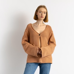 May include: A woman wearing a light brown knitted cardigan with a single button closure and a v-neck. She is wearing blue jeans.