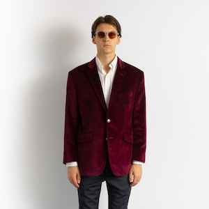 Puede incluir: Una blazer de terciopelo burdeos con cierre de dos botones, bolsillo en el pecho y dos bolsillos delanteros. La blazer se lleva sobre una camisa blanca y se combina con pantalones oscuros. El modelo lleva gafas de sol.