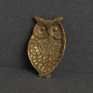 May include: A brass trinket dish shaped like an owl with detailed embossed feather patterns, prominent circular eyes, and pointed ear tufts. The metal surface has a warm, metallic gold tone with a polished finish and a concave center.