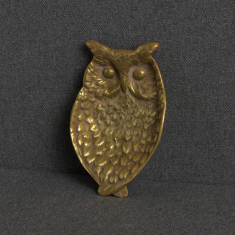 May include: A brass trinket dish shaped like an owl with detailed embossed feather patterns, prominent circular eyes, and pointed ear tufts. The metal surface has a warm, metallic gold tone with a polished finish and a concave center.