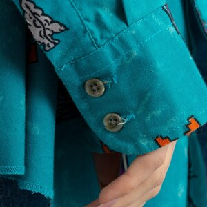 May include: A turquoise blue button-down shirt with a colorful abstract print. The shirt has two white buttons on the cuff.