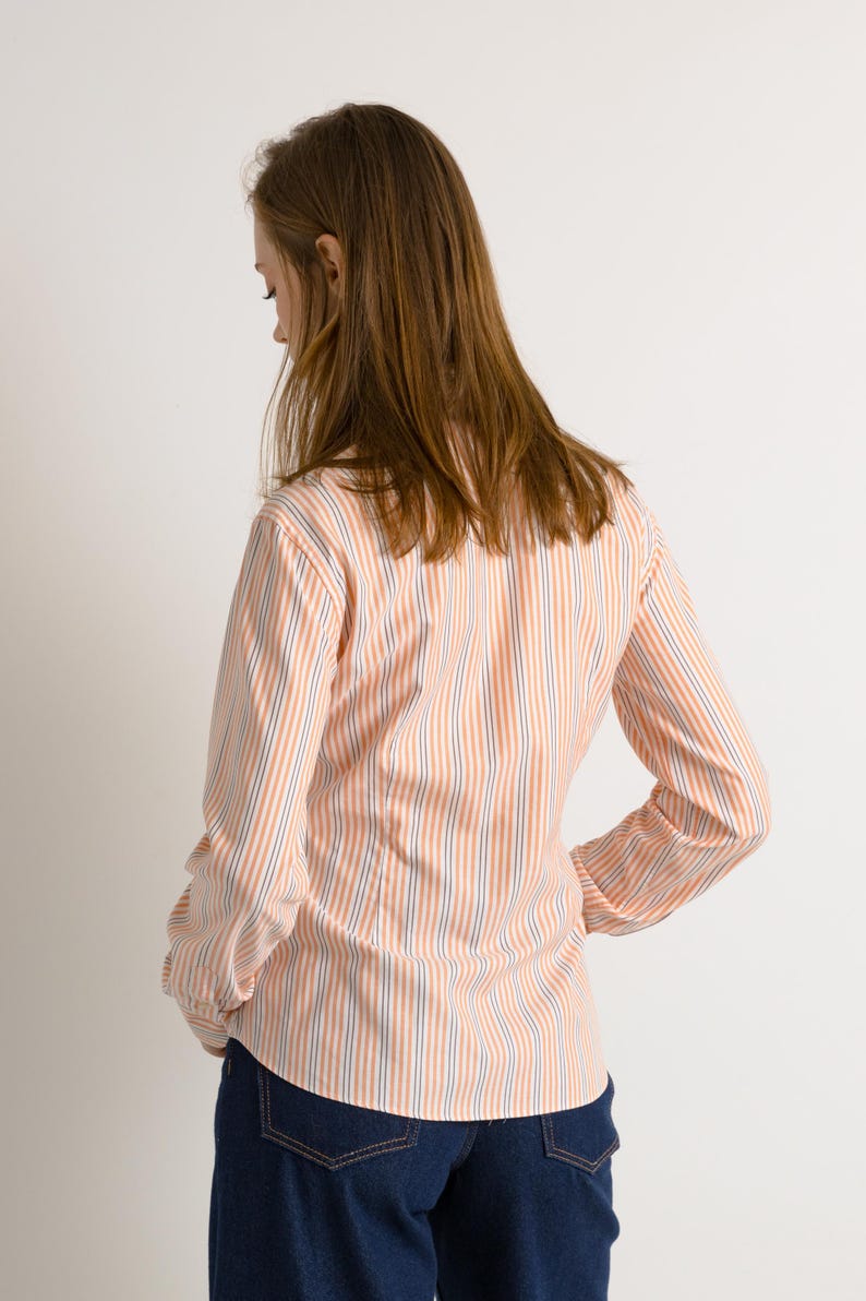Puede incluir: Una mujer lleva una camisa de manga larga a rayas con botones en tonos melocot&oacute;n y azul. La camisa tiene un corte holgado y est&aacute; metida en unos vaqueros azul oscuro.