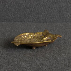 May include: A small, gold-toned brass dish shaped like a leaf with a textured surface featuring embossed grape clusters and foliage. The dish is supported by three small, rounded feet, providing a slightly elevated stance for this decorative metal tray.