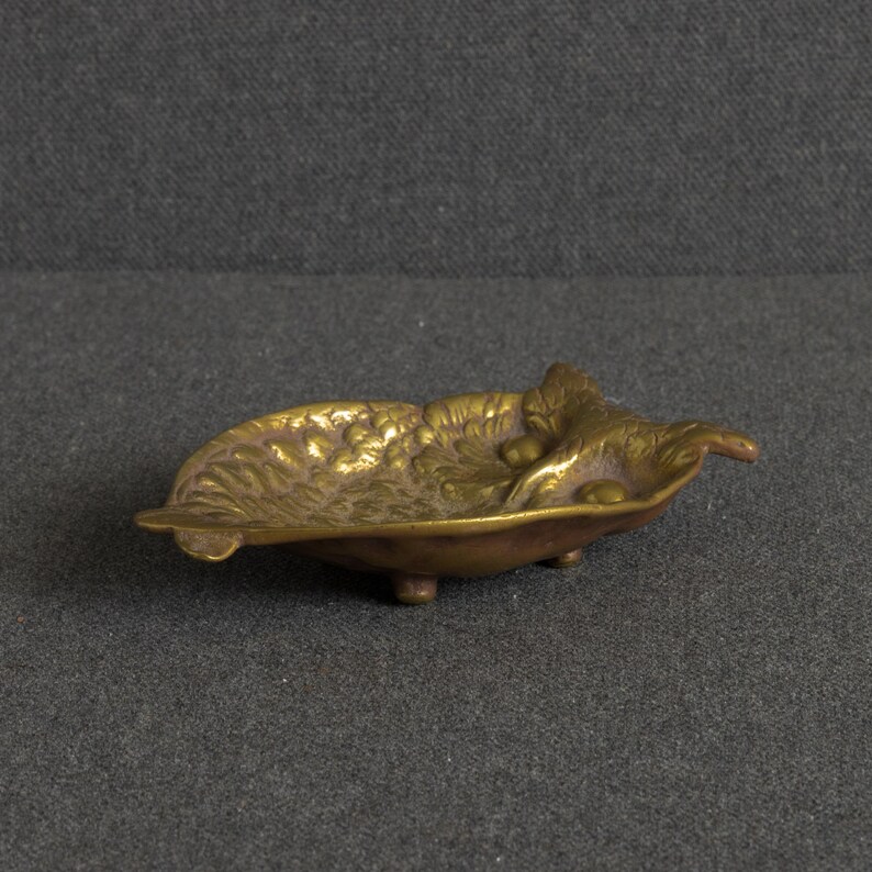 May include: A small, gold-toned brass dish shaped like a leaf with a textured surface featuring embossed grape clusters and foliage. The dish is supported by three small, rounded feet, providing a slightly elevated stance for this decorative metal tray.