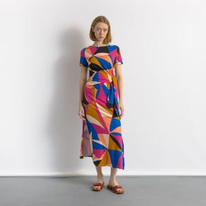 May include: A colorful geometric print dress with short sleeves and a wrap-style waist. The dress features a vibrant pattern of pink, blue, orange, and black shapes. The model is wearing brown sandals. The dress is knee-length.