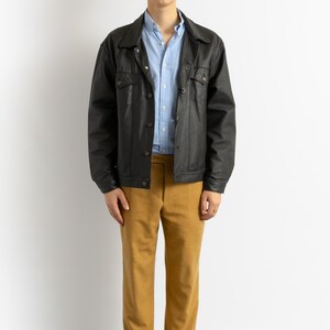 May include: A man wearing a black leather jacket over a light blue button-down shirt, brown pants, and black shoes.