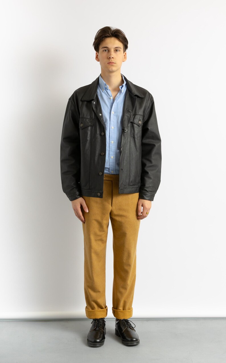 May include: A man wearing a black leather jacket over a light blue button-down shirt, brown pants, and black shoes.