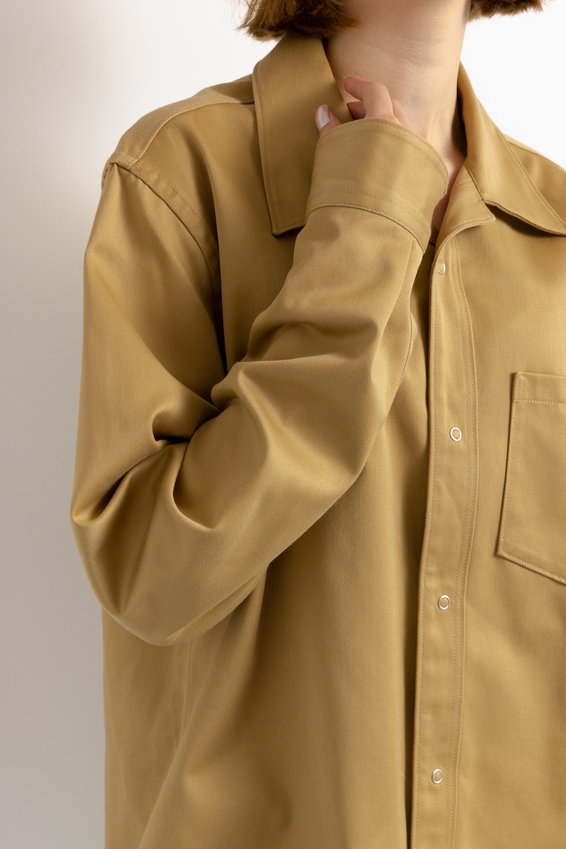 May include: A long-sleeved, tan button-down shirt with a single chest pocket and a relaxed fit.