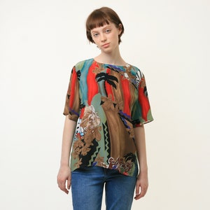May include: A woman wearing a short-sleeved, multi-colored blouse with an abstract print featuring palm trees, geometric shapes, and floral designs. The blouse has a brown, red, blue, green, and black color scheme. She is wearing blue jeans.