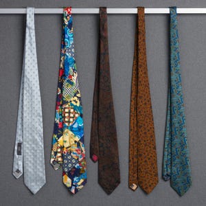 Puede incluir: Cinco corbatas colgadas de una barra plateada sobre un fondo gris. Las corbatas varían en color y patrón, incluyendo una corbata azul claro con lunares, una corbata floral de patchwork, una corbata marrón con estampado de cachemira, una corbata marrón con lunares y una corbata turquesa con estampado de cachemira.