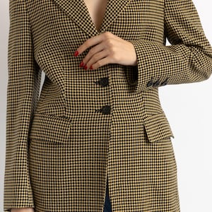 May include: A houndstooth blazer in black and tan. The blazer has a notched lapel, two front pockets, and three buttons on each cuff. The blazer is buttoned at the waist. The model is wearing blue jeans.