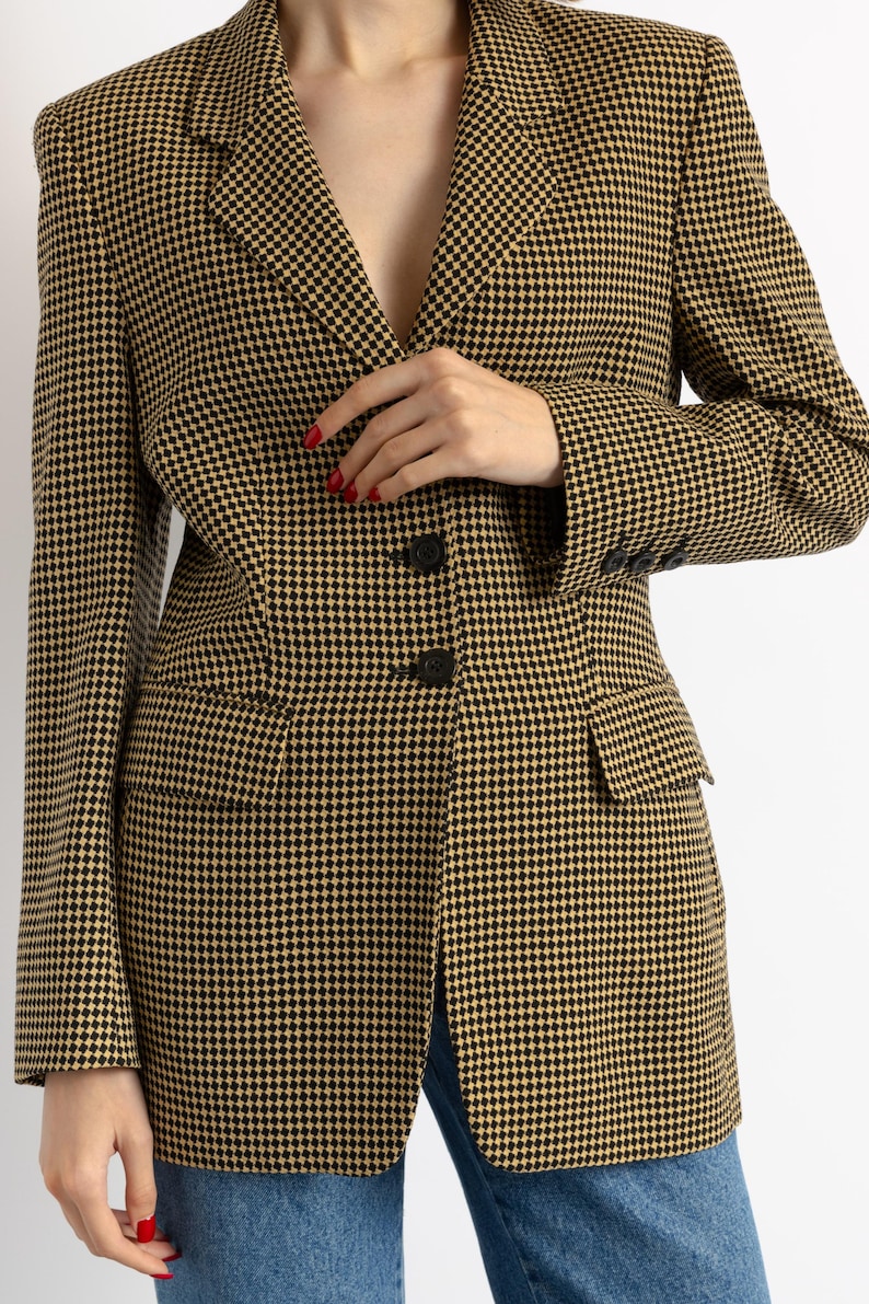 May include: A houndstooth blazer in black and tan. The blazer has a notched lapel, two front pockets, and three buttons on each cuff. The blazer is buttoned at the waist. The model is wearing blue jeans.