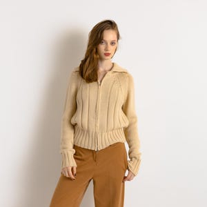 May include: A woman wearing a beige knitted cardigan with a zipper closure and brown pants.