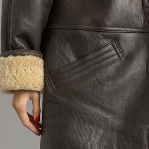 May include: Close-up of a brown leather coat with a shearling-lined cuff. The coat features a pocket and a button detail. The leather has a textured appearance.