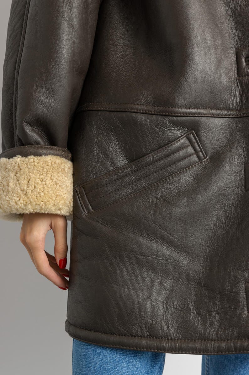 May include: Close-up of a brown leather coat with a shearling-lined cuff. The coat features a pocket and a button detail. The leather has a textured appearance.