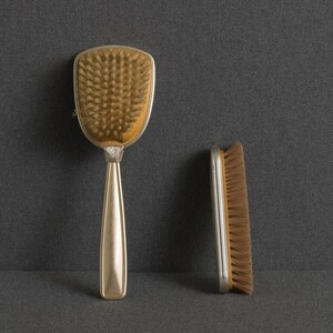 May include: A hairbrush with a metallic gold handle and a wide, rounded head featuring dense, light brown bristles, accompanied by a rectangular clothes brush with a metallic gold frame and matching light brown bristles.