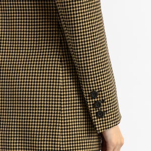 May include: A houndstooth blazer in black and tan. The blazer features a classic pattern and three black buttons on the sleeve. The fabric has a textured appearance. The blazer is paired with blue jeans.