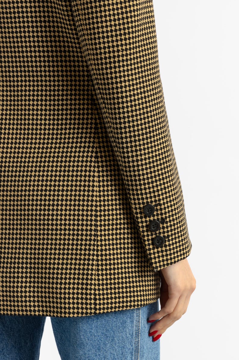 May include: A houndstooth blazer in black and tan. The blazer features a classic pattern and three black buttons on the sleeve. The fabric has a textured appearance. The blazer is paired with blue jeans.