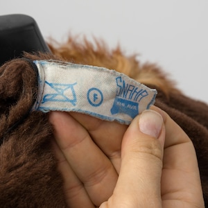 May include: Close-up of a brown faux fur garment with a light blue label. The label has the text "SNAHA" and other symbols. The garment has a dark brown color and a soft texture.