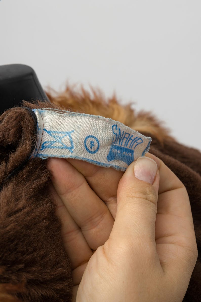 May include: Close-up of a brown faux fur garment with a light blue label. The label has the text "SNAHA" and other symbols. The garment has a dark brown color and a soft texture.