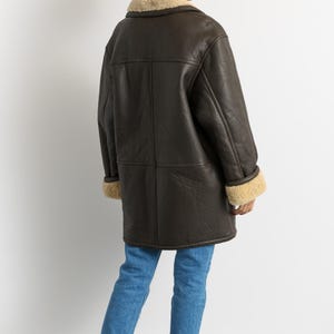 May include: A dark brown shearling coat with a cream-colored shearling collar and cuffs. The coat is worn over blue jeans and red boots. The coat is a long, warm style, perfect for cold weather.