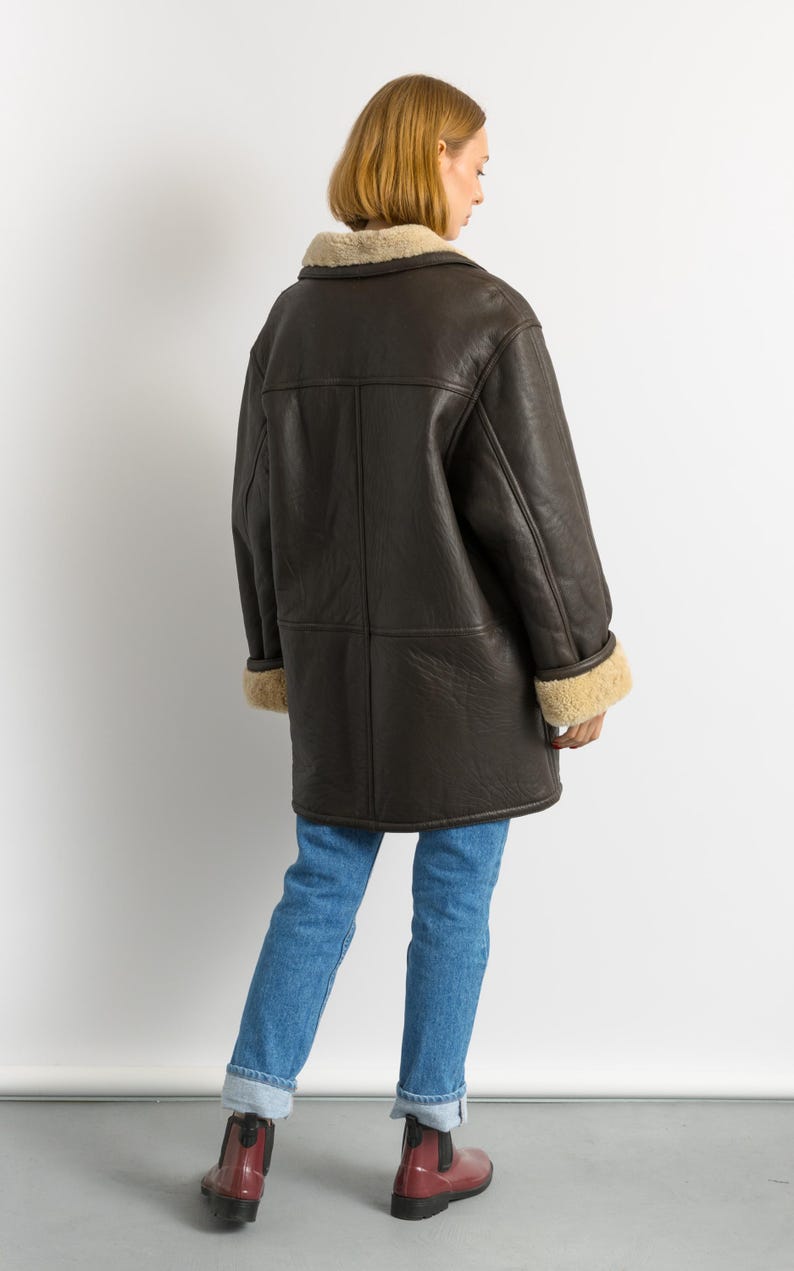 May include: A dark brown shearling coat with a cream-colored shearling collar and cuffs. The coat is worn over blue jeans and red boots. The coat is a long, warm style, perfect for cold weather.