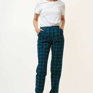 May include: A woman wearing a white t-shirt and green and blue plaid pants. She is standing with her hands in her pockets.