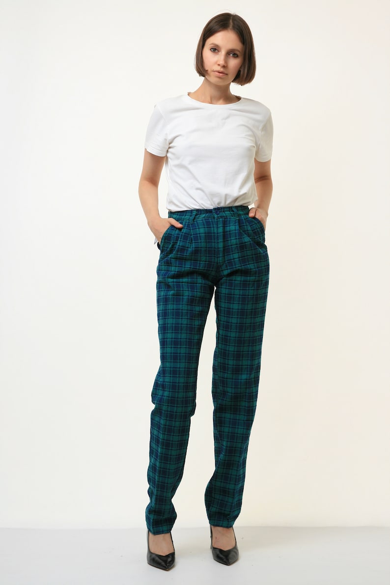 May include: A woman wearing a white t-shirt and green and blue plaid pants. She is standing with her hands in her pockets.