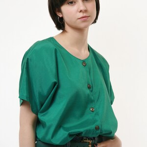 Puede incluir: Una mujer con una camisa verde de botones con mangas cortas y un cintur&oacute;n verde oscuro. Lleva pantalones vaqueros azules y tiene la mano en el bolsillo.