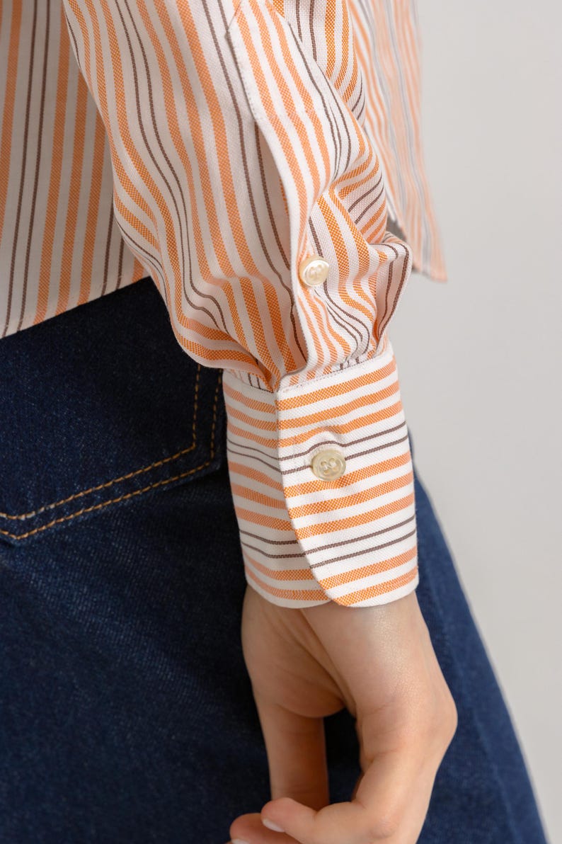 Puede incluir: Primer plano de una camisa de manga larga a rayas blancas y naranjas con rayas marrones. La camisa tiene un pu&ntilde;o con bot&oacute;n y un bot&oacute;n en la manga.