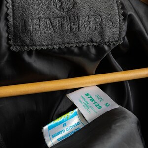 May include: Close-up of a black leather jacket with a label that reads "LEATHERS" and a size tag that says "SIZE M 87B125 GENUINE LEATHER 100% ACETATE". The tag also says "WE RECOMMEND SPECIALIST CLEANING BY".