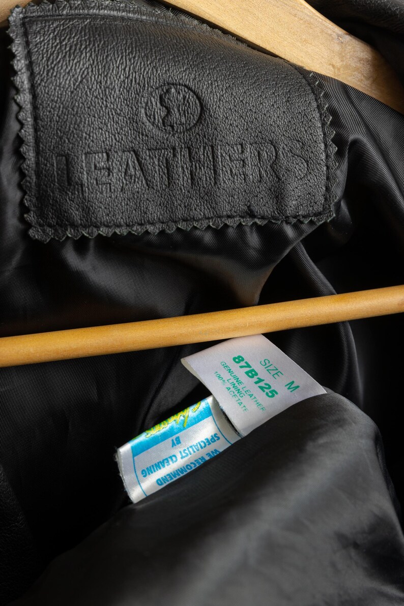 May include: Close-up of a black leather jacket with a label that reads "LEATHERS" and a size tag that says "SIZE M 87B125 GENUINE LEATHER 100% ACETATE". The tag also says "WE RECOMMEND SPECIALIST CLEANING BY".