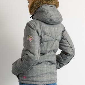 Puede incluir: Chaqueta de invierno acolchada gris con estampado de cuadros y capucha con forro de piel. La chaqueta tiene un peque&ntilde;o parche floral en la manga izquierda y un logotipo rosa cerca del dobladillo. La persona lleva vaqueros azules.